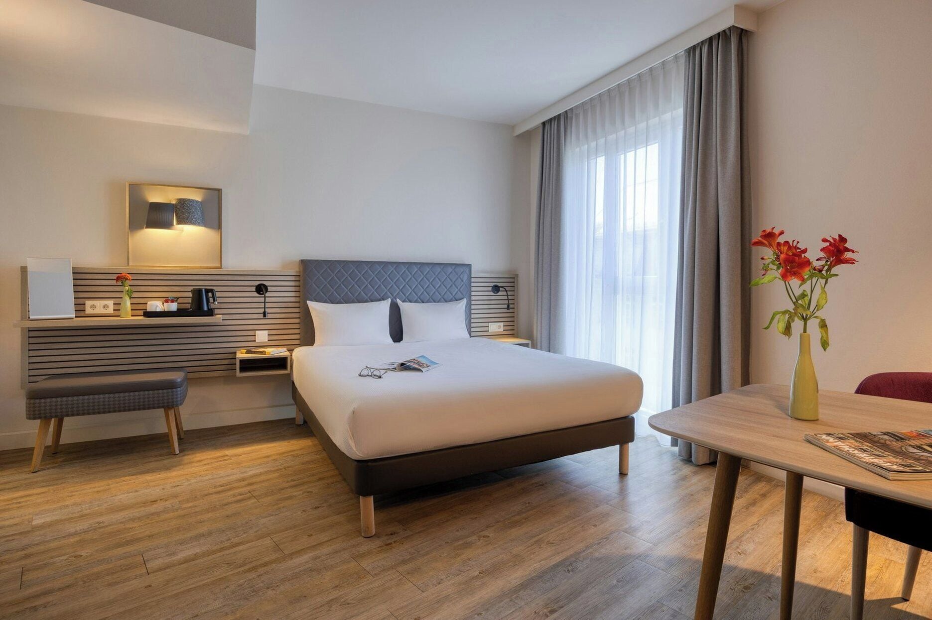 A hotel room with a bed, bedside table, desk, chair, and a view of the outdoors.