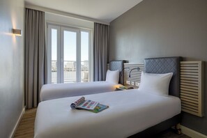 A hotel room with two beds, a large window with curtains, a headboard with a light fixture, and a view of the cityscape. - Citadines Arnulfpark Munich (Munich)