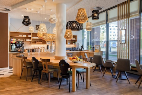 Ein modernes Café mit einem zentralen Holztisch, mehreren Stühlen, hängenden Pendelleuchten und einem großen Fenster mit Blick auf die Straße.. Citadines Arnulfpark Munich