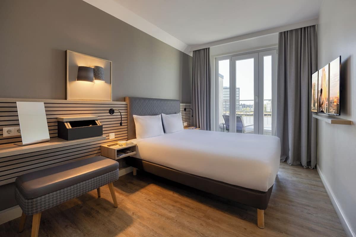 A hotel room with a bed, bedside table, lamp, and a view of the city through large windows.