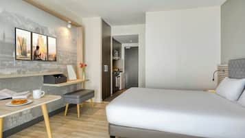 A modern hotel room with a large bed, a desk with a chair, a small kitchenette, and a view of the city.