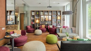 A modern living room with a large bookshelf, comfortable seating, and a large window. - Citadines Arnulfpark Munich (Munich)