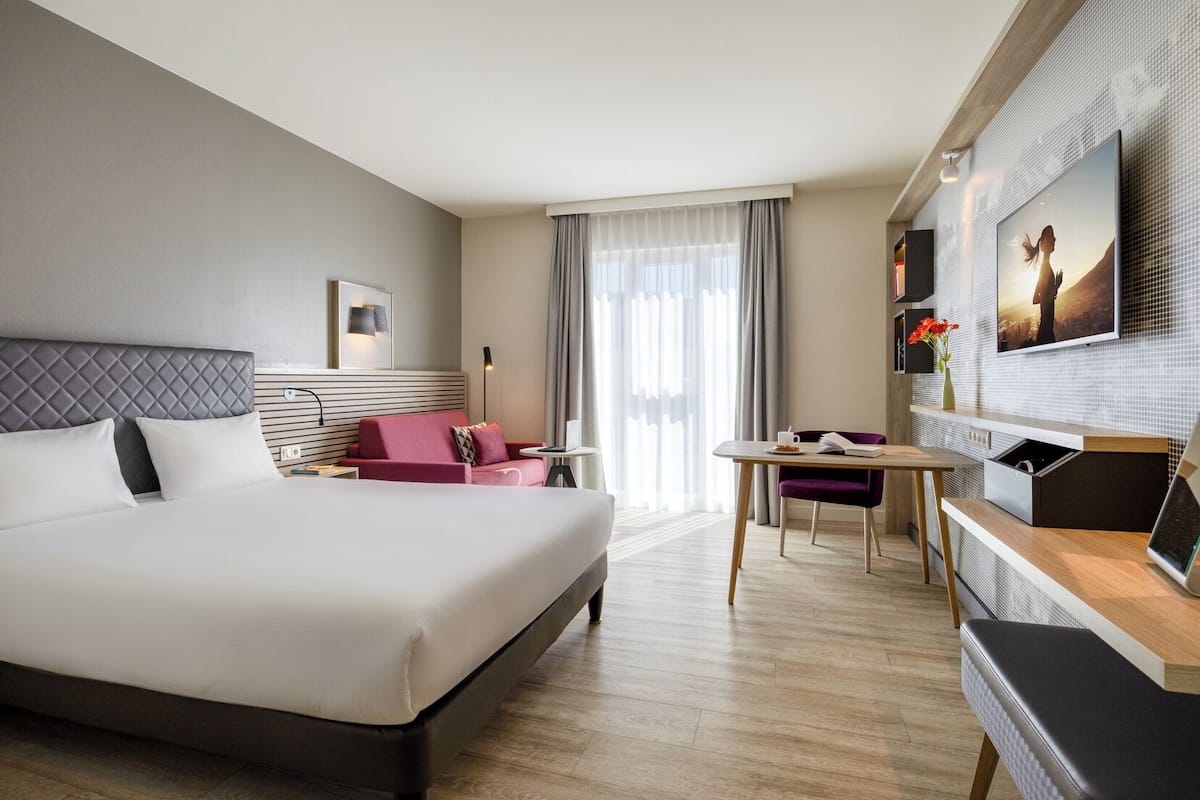 A hotel room with a large bed, a sofa, a dining table, and a TV mounted on the wall.