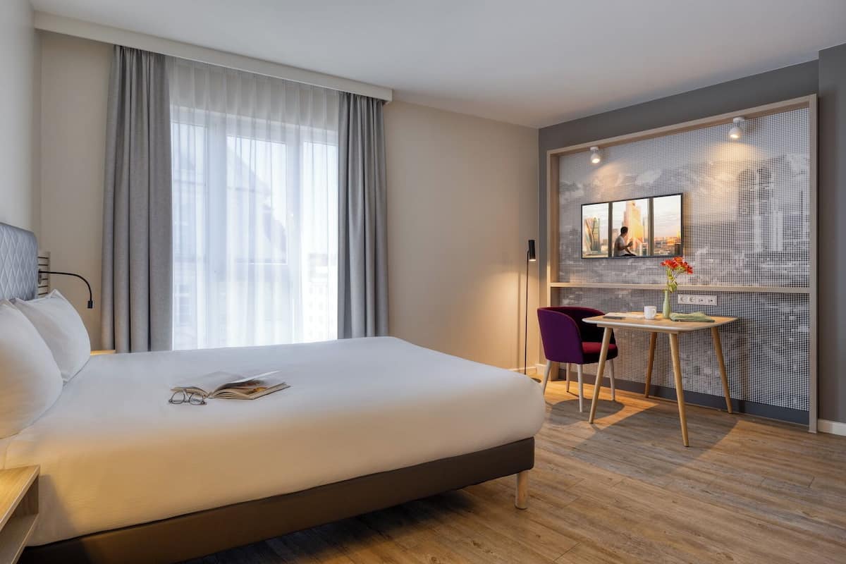 A hotel room with a large bed, a small table with a chair, a window with curtains, and a wall-mounted TV.