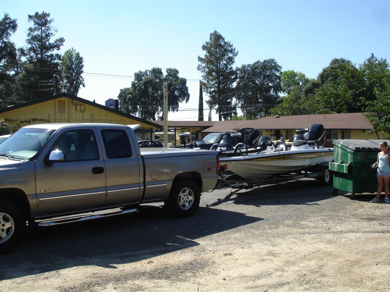 A pickup truck towing a boat and a green dumpster in a parking lot with buildings in the background.