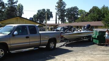 A pickup truck towing a boat and a green dumpster in a parking lot with buildings in the background.