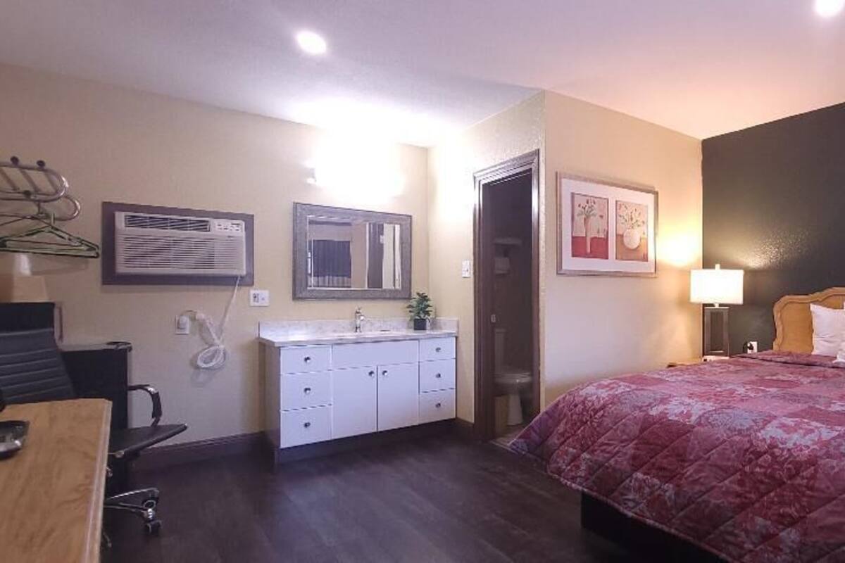A hotel room with a bed, bedside table, lamp, and a desk with a chair. There is a door leading to another room and a wall-mounted air conditioner.