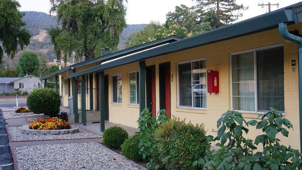 A motel with a yellow exterior, green trim, and a red door. It has a gravel driveway and is surrounded by greenery.