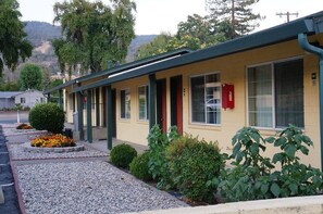 A motel with a yellow exterior, green trim, and a red door. It has a gravel driveway and is surrounded by greenery.