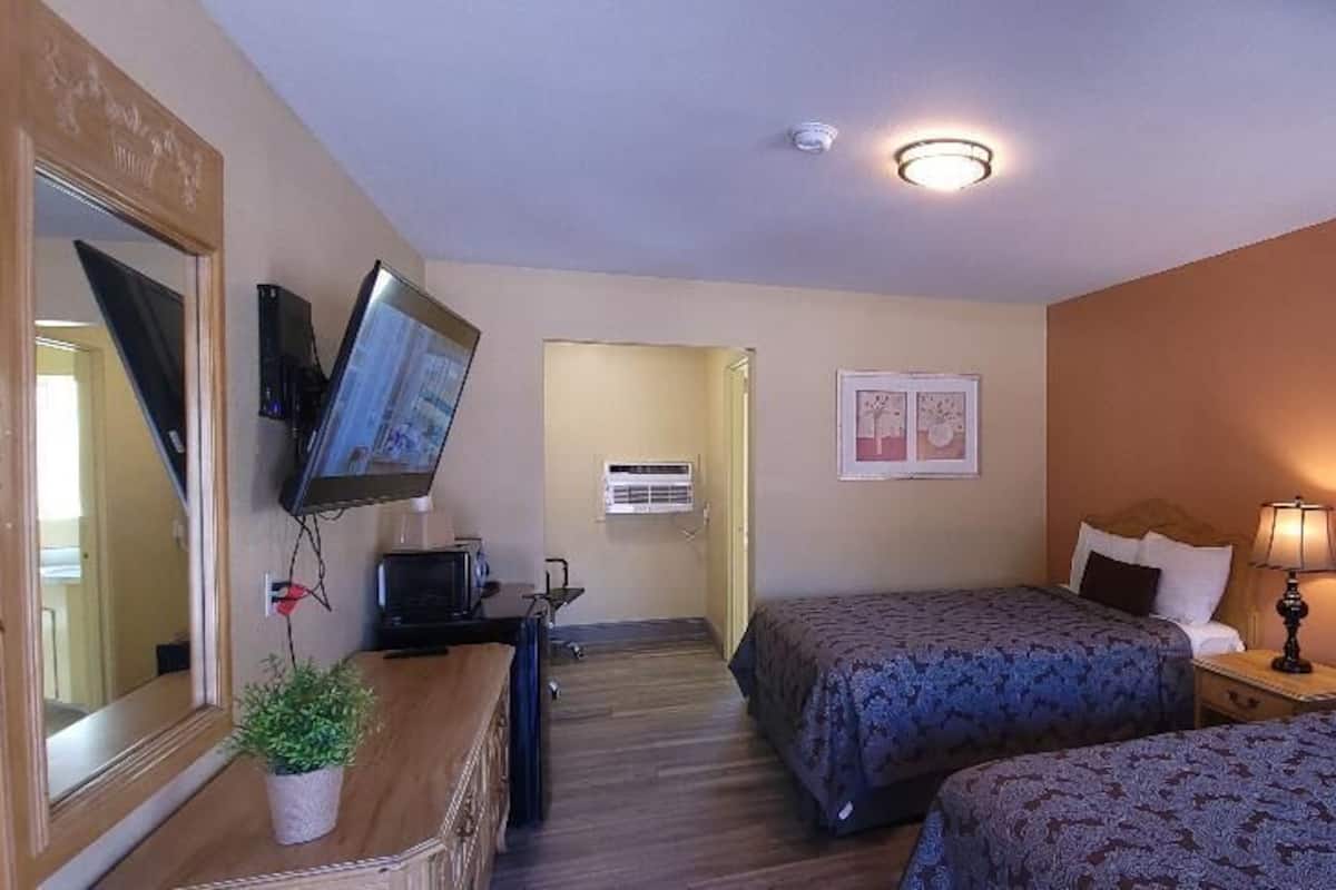 A hotel room with two beds, a TV mounted on the wall, a desk with a computer, and a potted plant.
