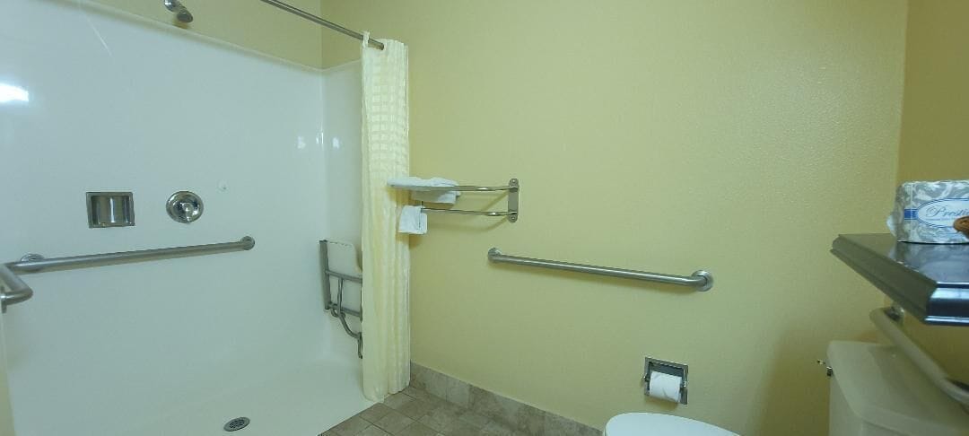 Un baño con ducha, barras de apoyo, inodoro y cubo de basura.