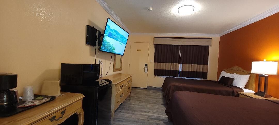 A hotel room with a TV mounted on the wall, a desk with a computer, a bed with a headboard, and a window with curtains.