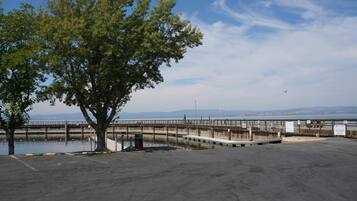 A waterfront parking area with a large tree, a wooden boardwalk, and a view of the water and distant hills.