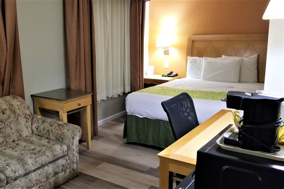 A hotel room with a floral sofa, a small table, a bed with a green bedspread, a nightstand with a lamp, and a coffee maker on the table.
