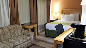 Habitación de hotel con un sofá floral, una mesita, una cama con colcha verde, una mesita de noche con lámpara y una cafetera sobre la mesita.