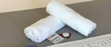 A neatly made bed with white linens, a pair of blue flip-flops, a towel, and a small bottle on a grey surface.
