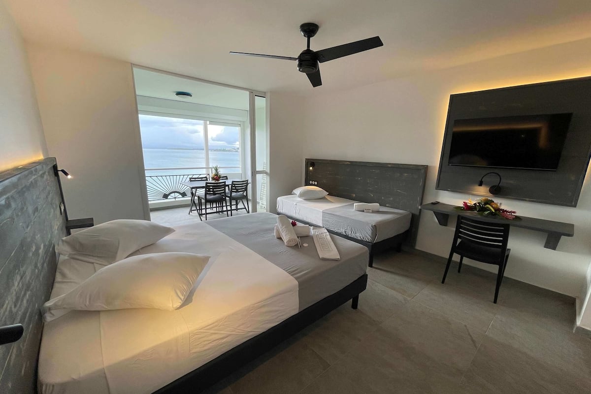 Habitación de hotel con dos camas, ventilador de techo, televisión y balcón con mesa y sillas.