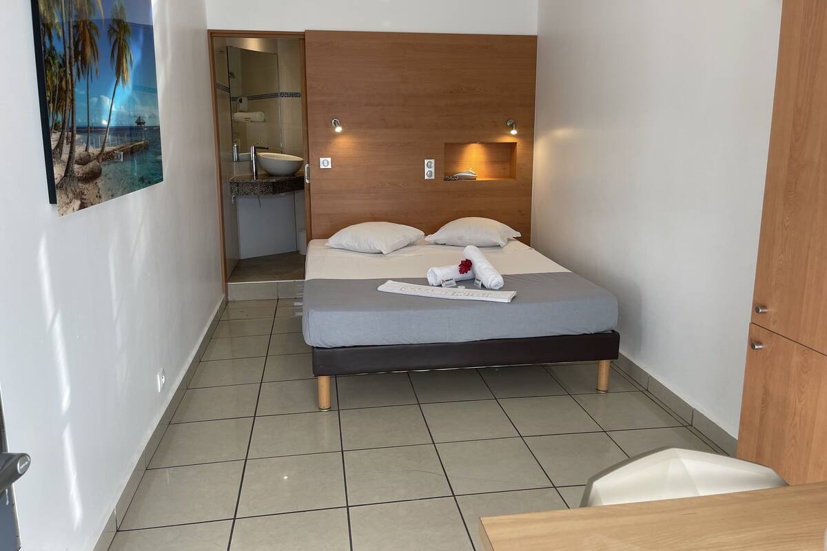 Una habitación de hotel moderna con cama, cabecera de madera, baño con lavabo e inodoro, y un cuadro con una playa.