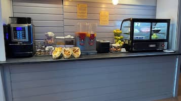 Un mostrador de desayuno autoservicio con una máquina de café, pan, bollería y una exhibición de frutas.