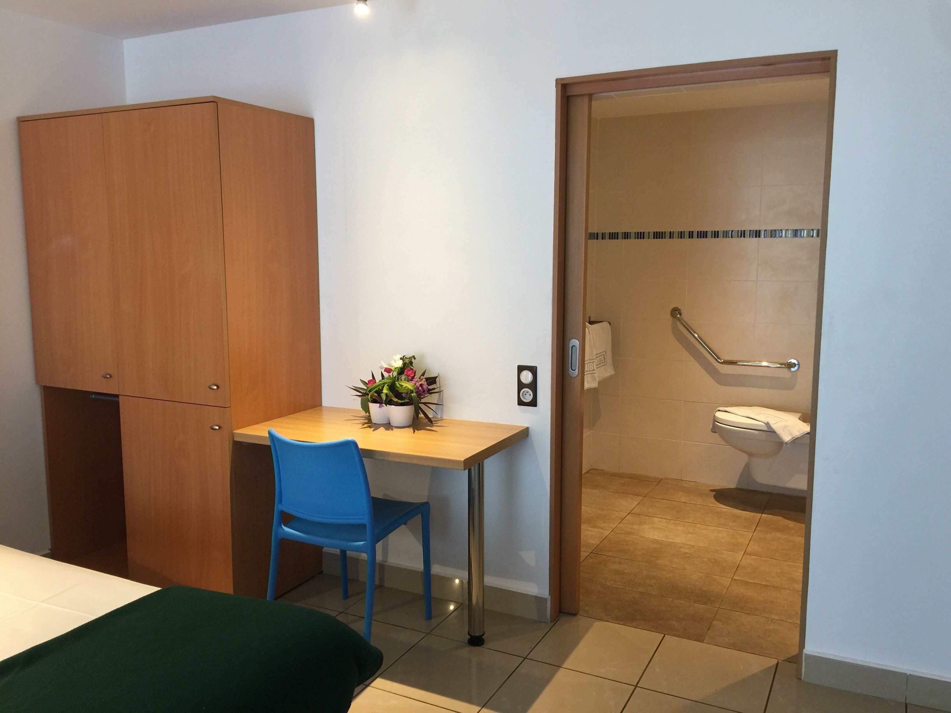 A small room with a wooden wardrobe, a blue chair, a small table with a potted plant, and a door leading to a bathroom with a toilet and a handrail.