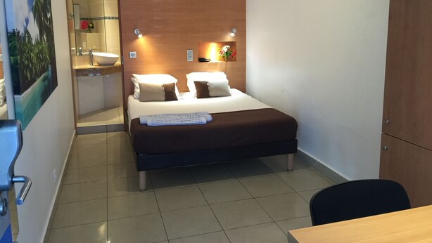 A hotel room with a bed, a desk, a chair, a bathroom with a sink and mirror, and a wall-mounted TV. - Karaibes Hotel (Le Gosier)
