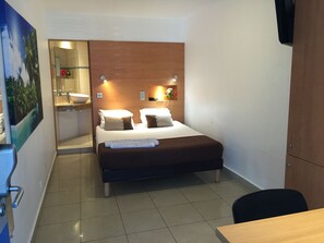 A hotel room with a bed, a desk, a chair, a bathroom with a sink and mirror, and a wall-mounted TV. - Karaibes Hotel (Le Gosier)