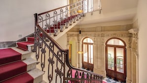 A grand staircase with ornate metal railings and red carpeted steps leading to a balcony area with arched doorways and a checkered floor.