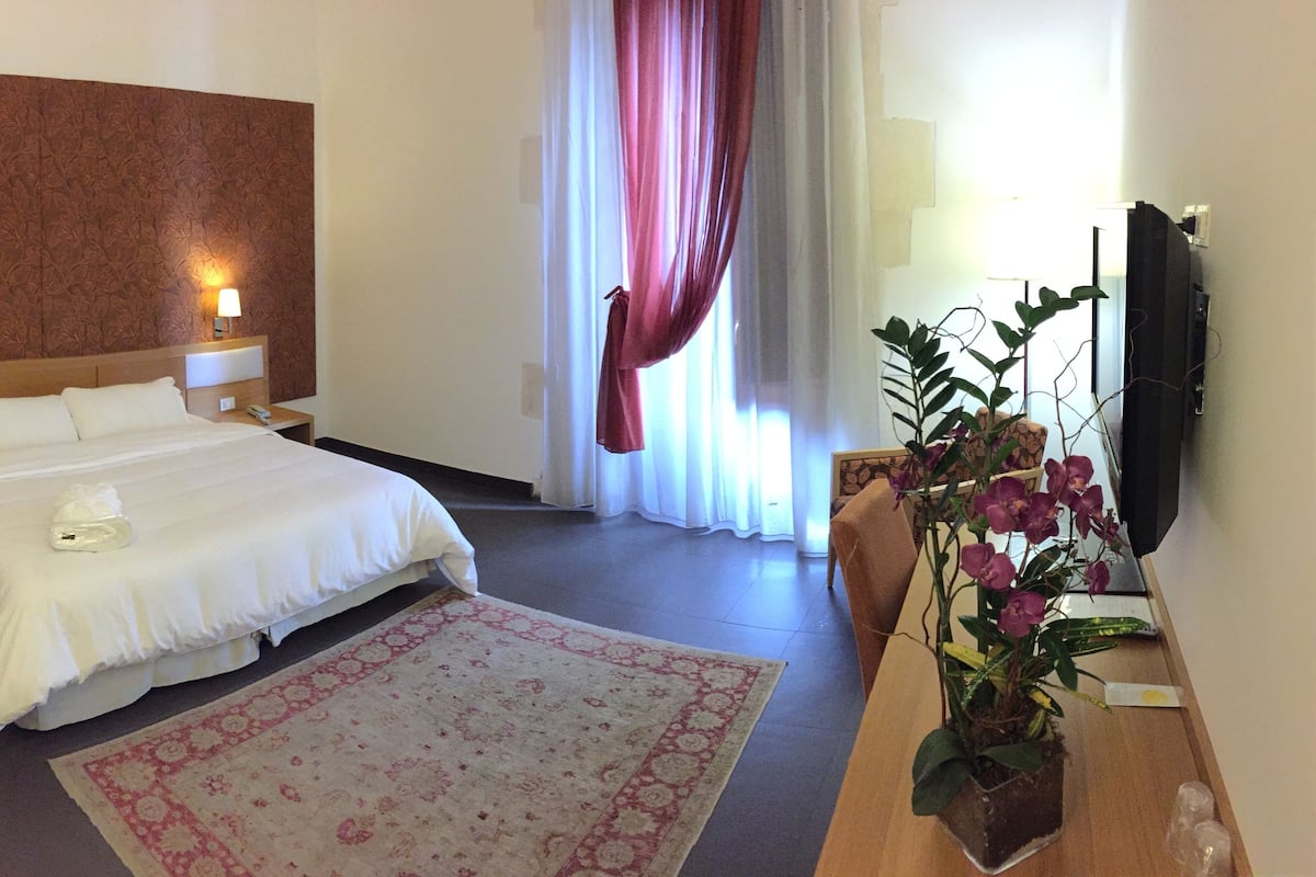 A hotel room with a large bed, two bedside tables with lamps, a wooden headboard, a large window with curtains, a potted plant, and a flat-screen TV.