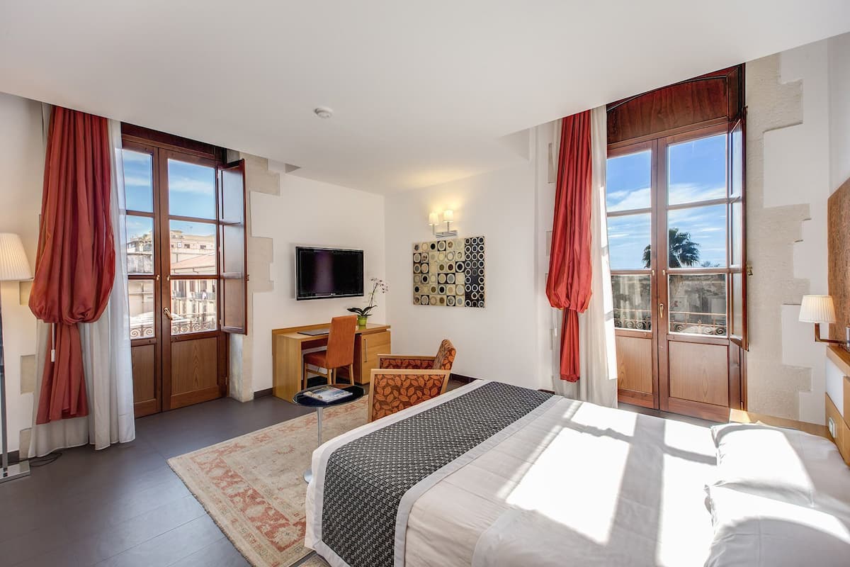 A hotel room with a bed, desk, chair, TV, and large windows with curtains.