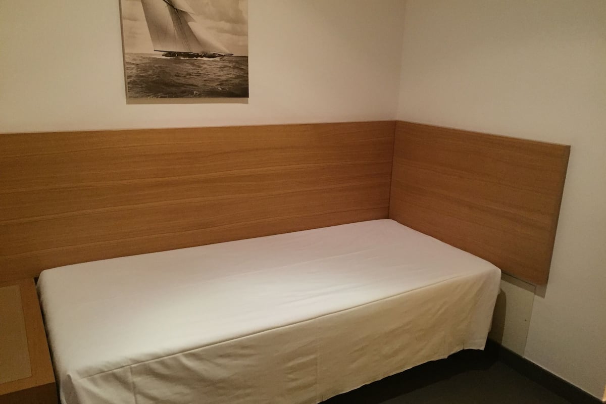A small, single-bed room with a wooden headboard, a white mattress, and a framed picture of a sailboat on the wall.
