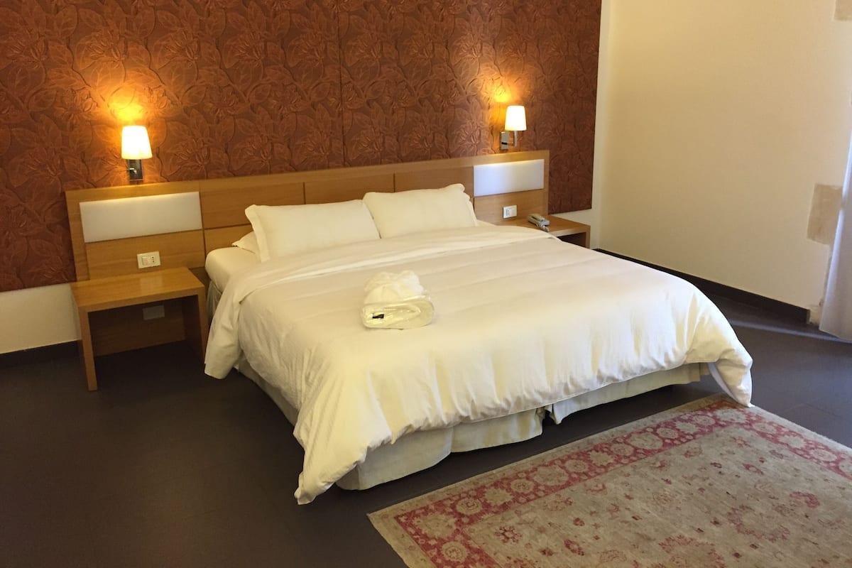 A hotel room with a large bed, two bedside tables, a nightstand, a lamp, and a patterned rug.