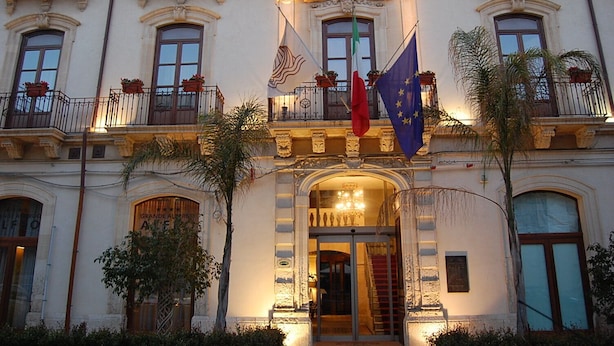 A historic building with ornate balconies, a grand entrance, and flags of the European Union and Italy.