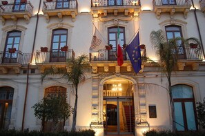 Un bâtiment historique doté de balcons richement décorés, d’une entrée imposante et arborant les drapeaux de l’Union européenne et de l’Italie.