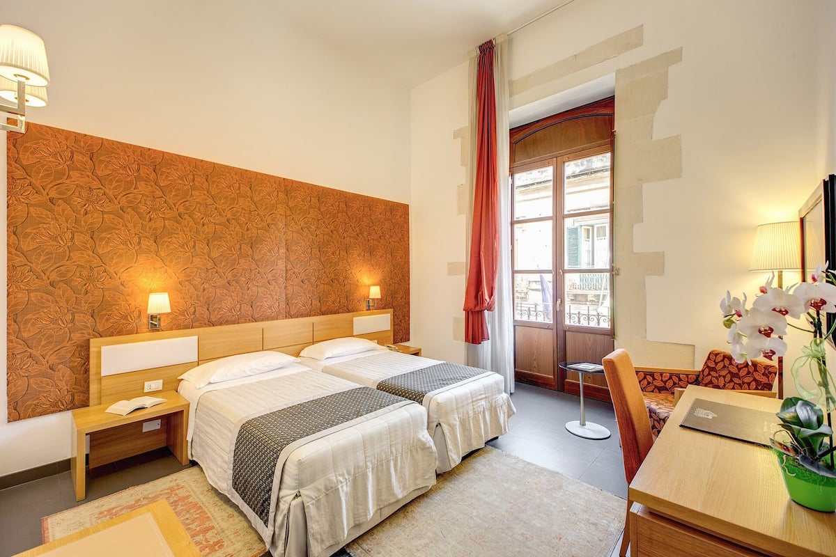 A hotel room with two beds, a desk, a chair, a window with a view, and a potted plant.