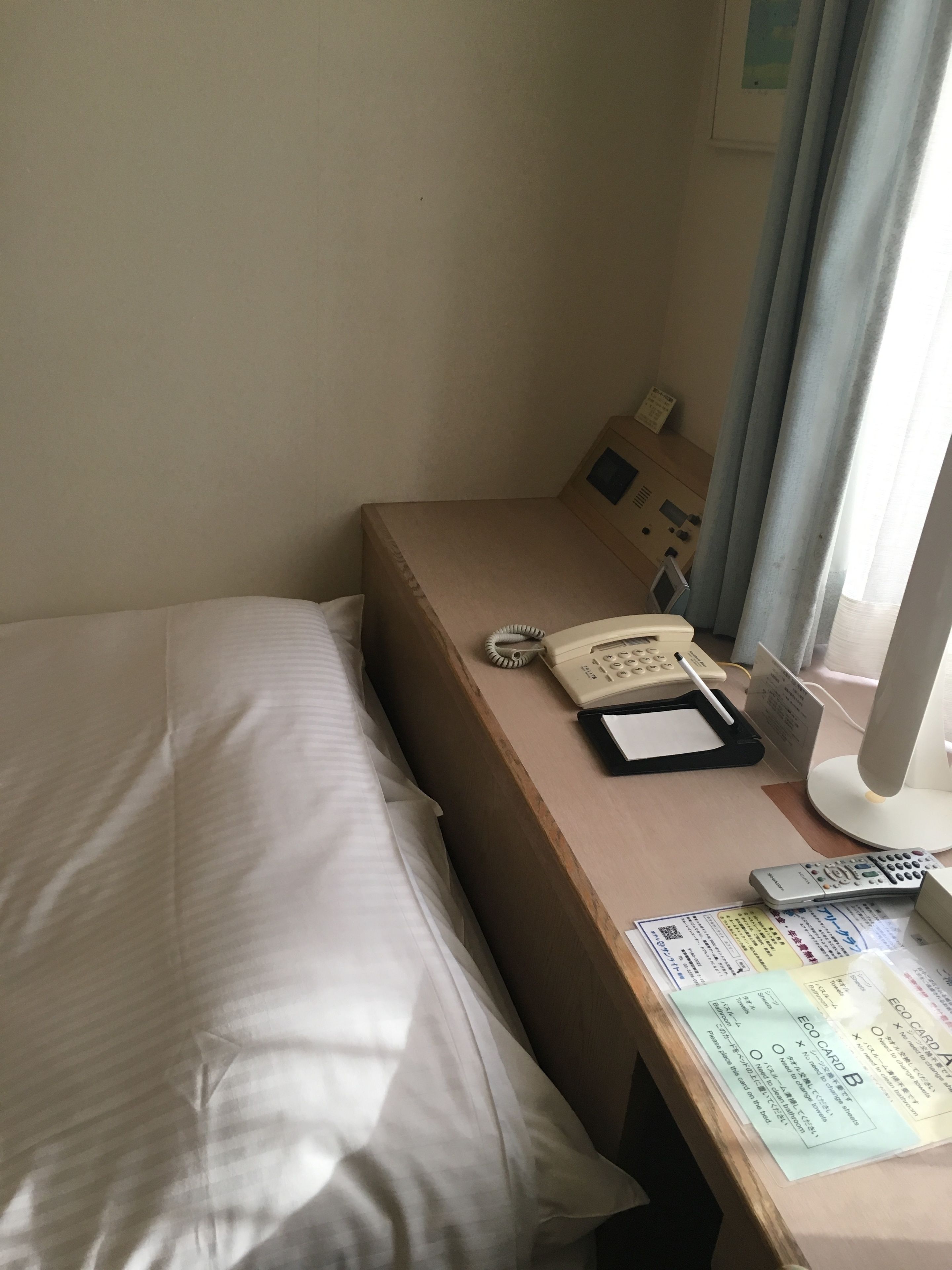 A hotel room with a bed, a bedside table with a telephone, a remote control, and a desk with a computer monitor and papers.