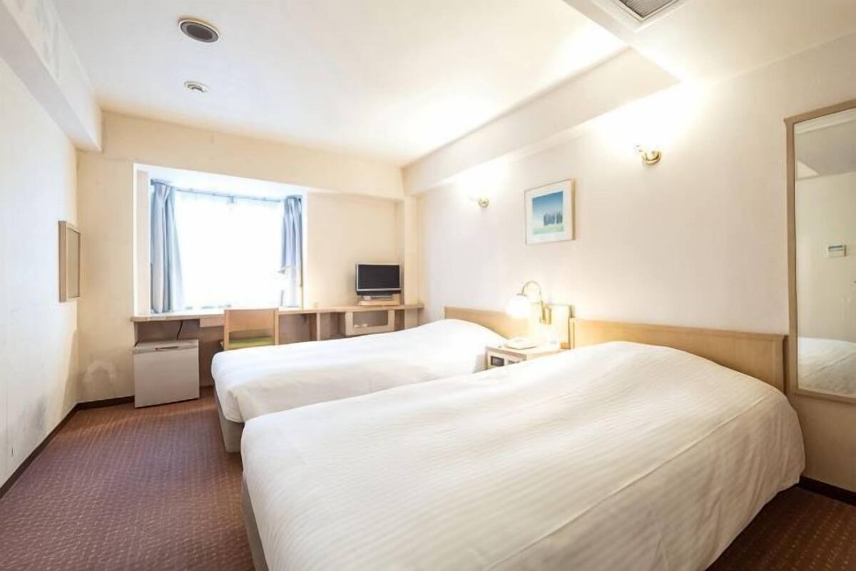 A hotel room with two beds, a desk, a television, and a window with curtains.