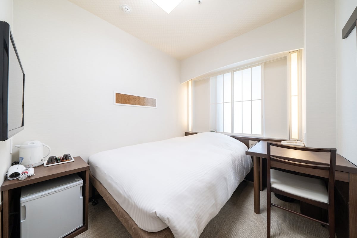 A hotel room with a bed, a desk, a chair, a small refrigerator, and a window with natural light.