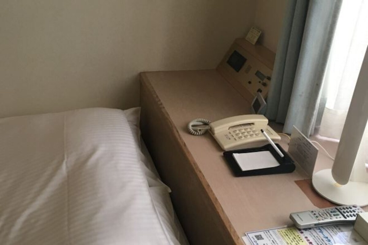 A hotel room with a bed, a bedside table with a telephone, a remote control, and a small desk with a computer monitor.