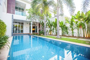 Et moderne poolområde med en klar, blå pool, omgivet af frodigt grønt og en toetagers bygning med store vinduer og en balkon.