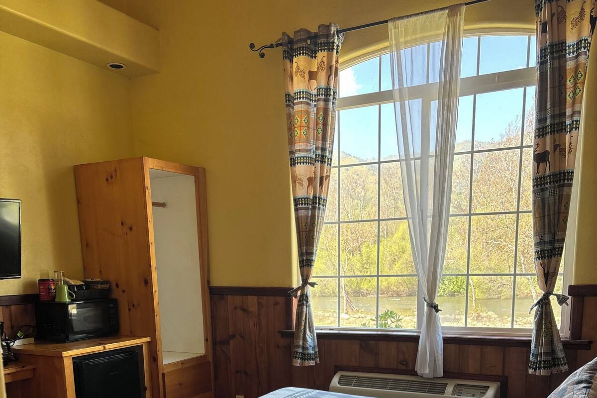 Un dormitorio con cama, armario de madera, una pequeña kitchenette y una ventana con cortinas.