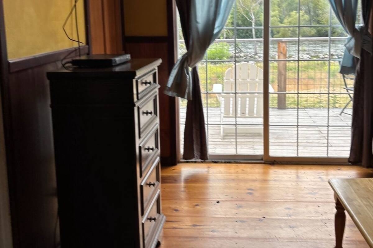 Una habitación con piso de madera, un aparador, un televisor montado en la pared y un ventanal con cortinas.