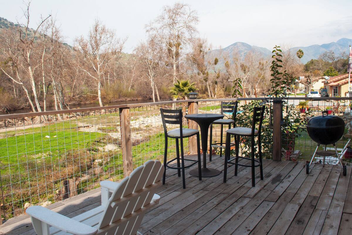 Una terraza de madera con mobiliario exterior, una parrilla y vistas a un espacio verde y montañas.