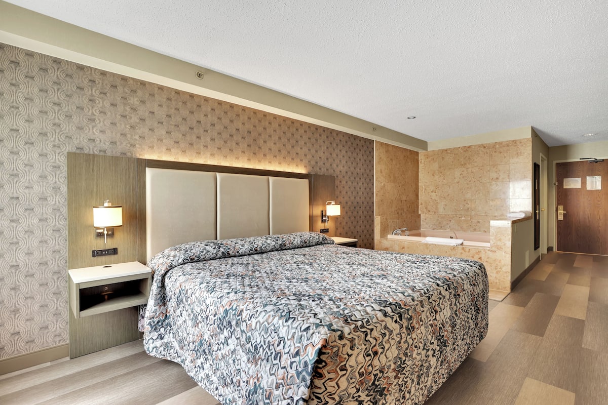 A hotel room with a large bed, a nightstand, a wall-mounted lamp, and a bathroom with a bathtub and sink.