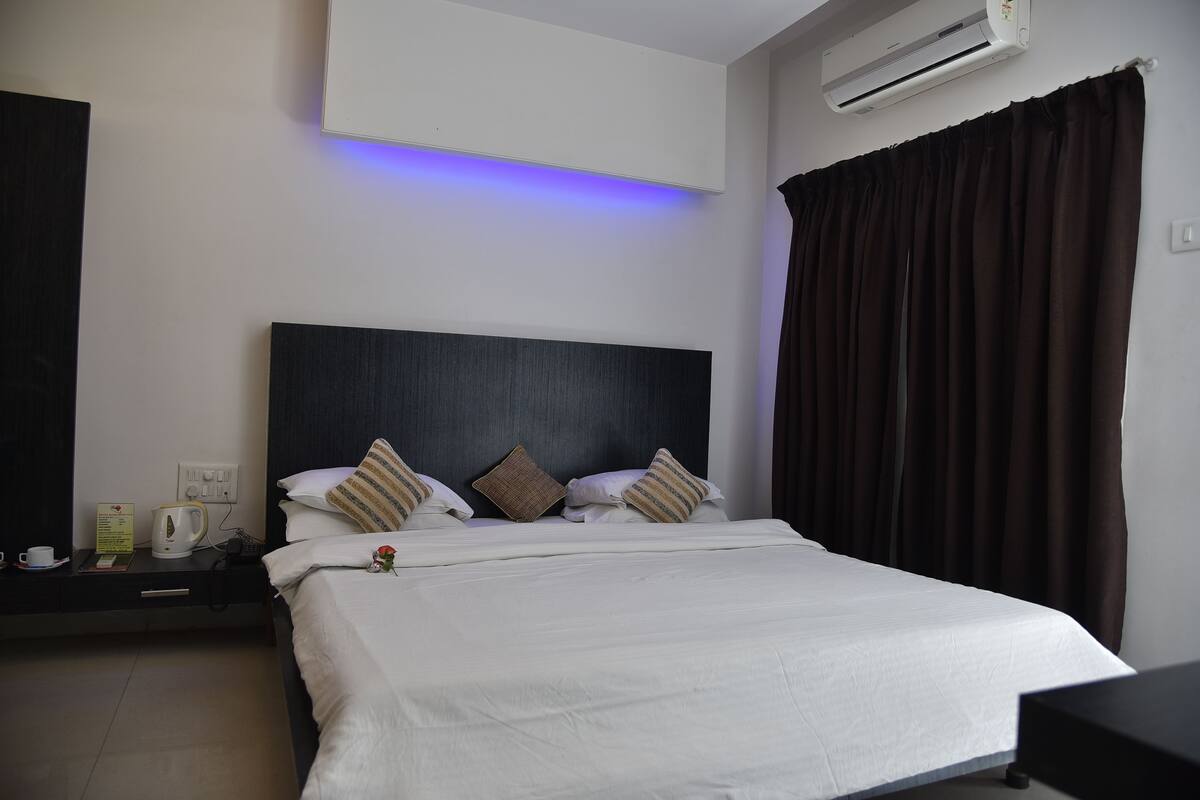 A neatly made bed with white linens, a dark headboard, and two pillows. There is a bedside table with a kettle, a cup, and a sign. The room has a modern design with a large window covered by dark curtains.