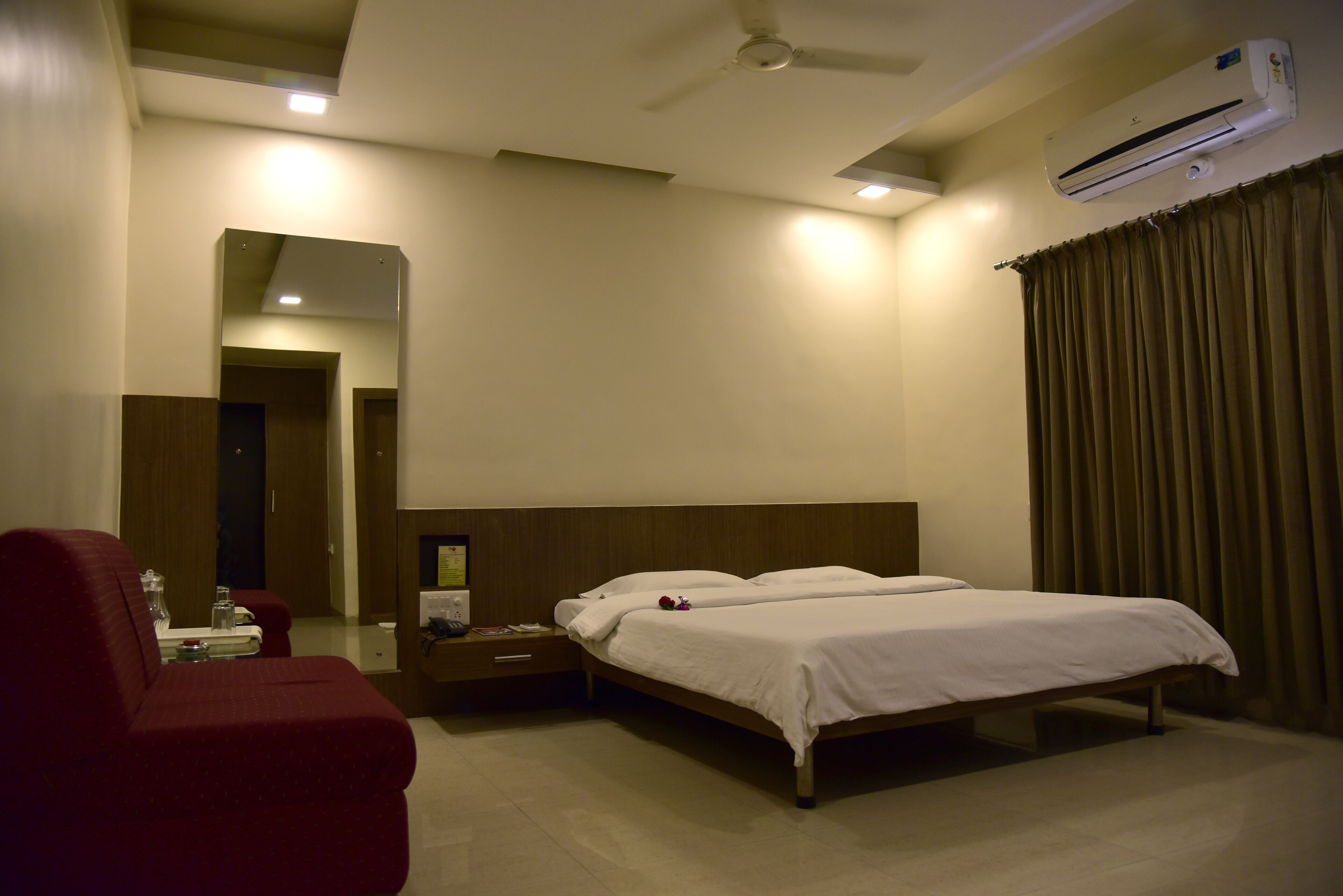 A hotel room with a bed, a red armchair, a nightstand, a mirror, and an air conditioning unit.