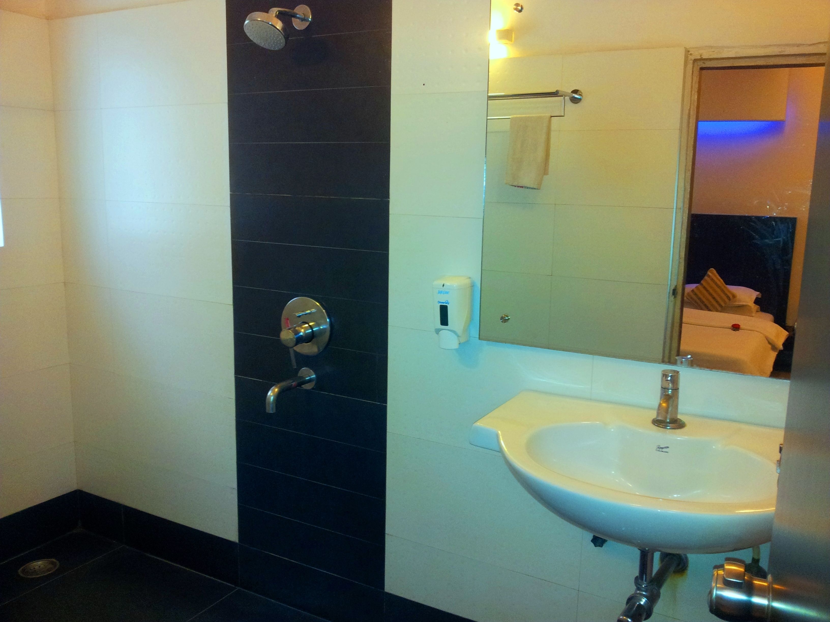 A bathroom with a black tiled shower wall, a white sink, and a mirror reflecting a bed with a towel.