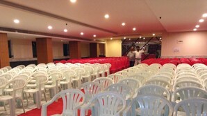 Rows of white plastic chairs arranged in a banquet hall with a red carpet, a stage with a backdrop, and a ceiling with recessed lighting.