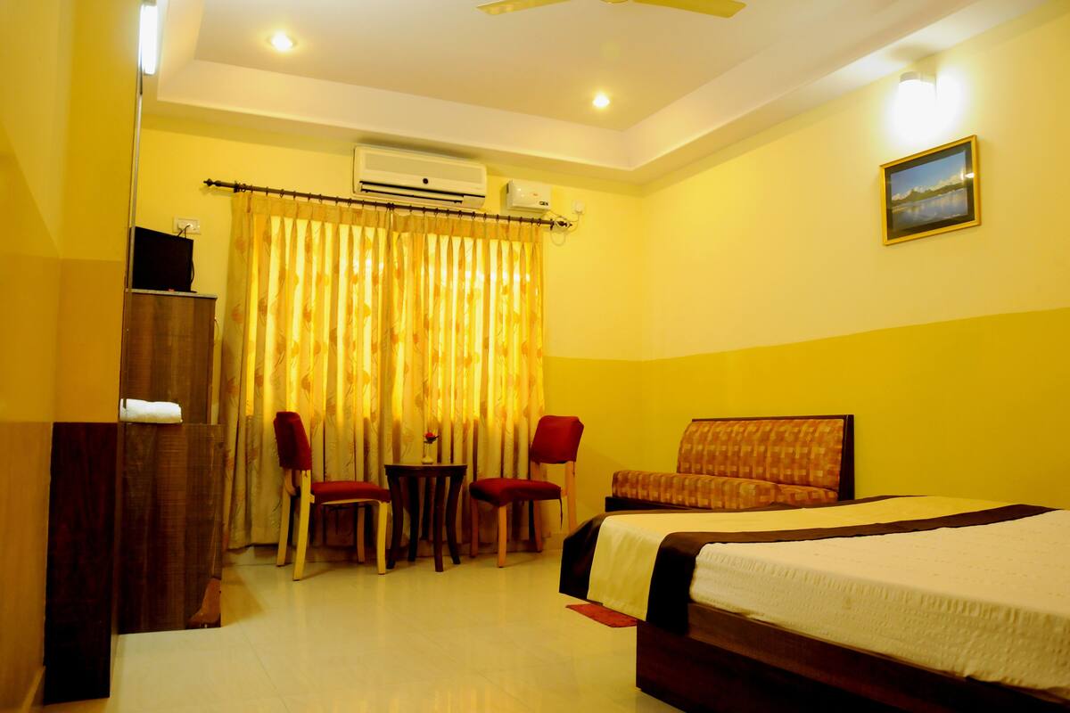 A hotel room with a bed, a desk, two chairs, a small table, an air conditioning unit, and a picture on the wall.