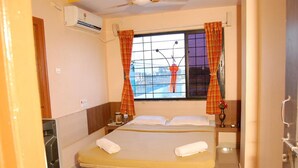 A bedroom with a bed, curtains, an air conditioning unit, a window with a view, and a bedside table with a lamp. - Hotel Utsav (Rahata)