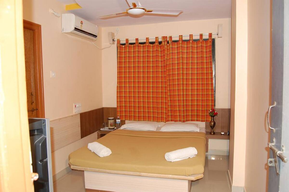 Une chambre d’hôtel comprenant un lit, un ventilateur de plafond, un appareil de climatisation, une table de chevet et une fenêtre avec des rideaux à carreaux.
